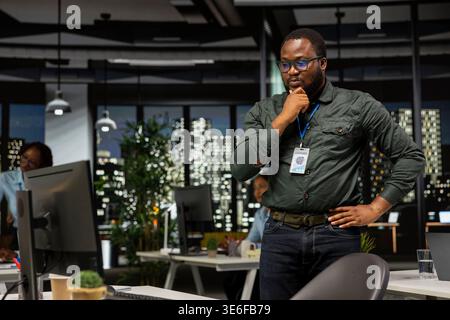 African american director reflecting logical solution and scratching his chin, works after hours at desk. Man engaging in critical thinking, evaluating each option for a different perspective. Stock Photo