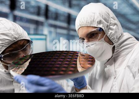Fabrication engineers checking silicon wafer hardware defects in industry 4.0 manufacturing plant. Specialists in bunny suits assessing smart factory semiconductor substrate yields, close up Stock Photo