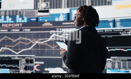 Woman using tablet to compare company KPIs data on digital videowall before making decisions. African american entrepreneur analyzing key performance indicators using mockup phone, camera B Stock Photo