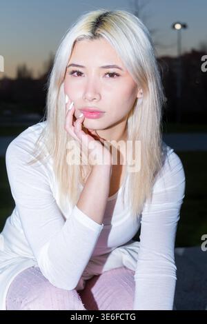 Close-up flash portrait of a Gen Z girl with platinum blonde hair and soft makeup, resting her chin on her hand outdoors at dusk, with bokeh city ligh Stock Photo
