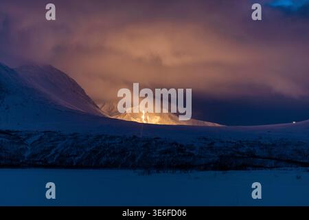 Dramatic winter night scene near Tromso, Norway, with snow-covered mountains and valley under heavy clouds, glowing light on the horizon in Arctic Lap Stock Photo