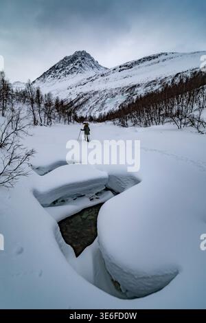 Lone snowshoer hikes across a pristine snowy valley toward rugged mountains near Tromso, Norway, in Arctic Lapland, beneath an overcast winter sky, ex Stock Photo