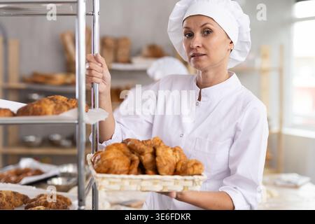 Portrait of old female baker working in bakehouse, holding tray with fresh bakery goods on kitchen Stock Photo