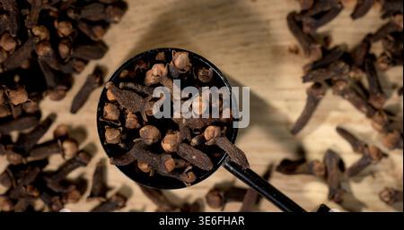 Cloves in black scoop on wooden surface with scattered cloves around it Stock Photo