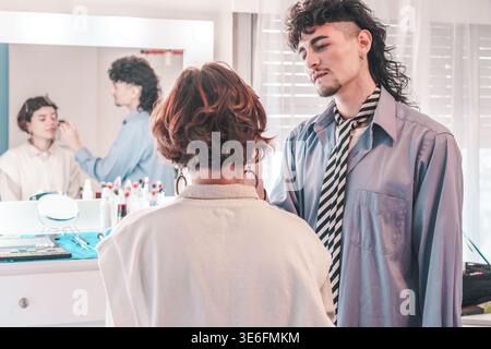 Makeup artist applies makeup on a model in a brightly lit dressing room, consulting with a stylist about the final look while another makeup artist wo Stock Photo