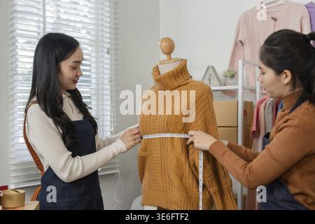 Two professional female designers meticulously measuring a cozy knitted sweater on a mannequin, working together in a trendy fashion studio Stock Photo