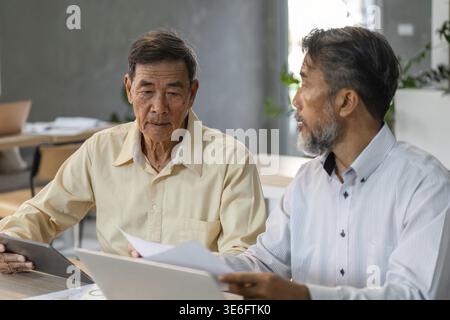 Two middle-aged businessmen sit in a contemporary office, engaging in a serious discussion about financial strategies, exchanging documents and ideas Stock Photo
