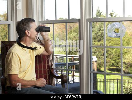 Senior man watching chickadee bird on birdfeeder through binoculars from chair Stock Photo