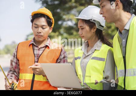 Three construction workers, including an engineer and a supervisor, are analyzing plans and discussing measurements on-site while ensuring safety with Stock Photo