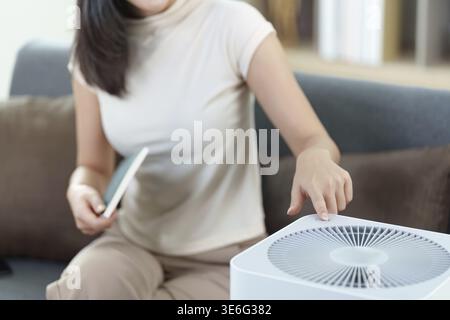 A woman interacts with a smart air purifier using her smartphone, showcasing the convenience of smart home technology in a cozy living space Stock Photo