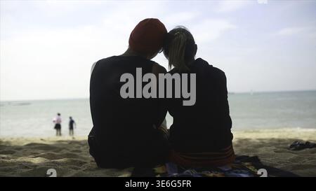 Rear view of Couple sitting on the sand by the sea and back nature of summer. Video. Happy Loving couple on a cliff view tropical beautiful crystal cl Stock Photo
