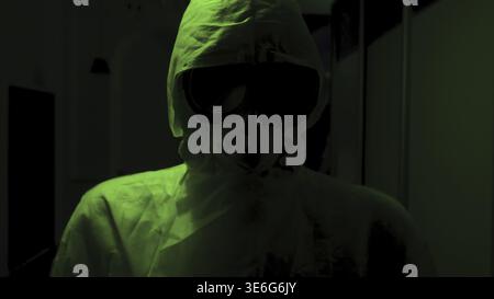 Man in protective suit and mask stands in dark. Stock footage. Creepy doctor in antiviral suit stands in dark corridor with green light. Dangerously h Stock Photo