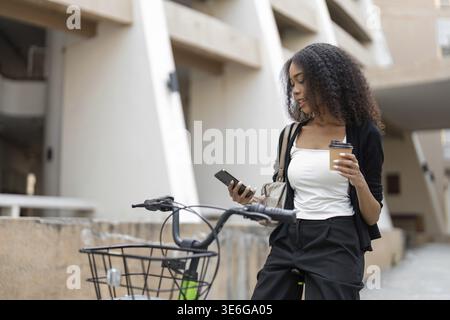 Pausing for a moment, a woman enjoys her coffee as she checks her phone, seamlessly blending urban life with sustainable commuting on her bicycle Stock Photo