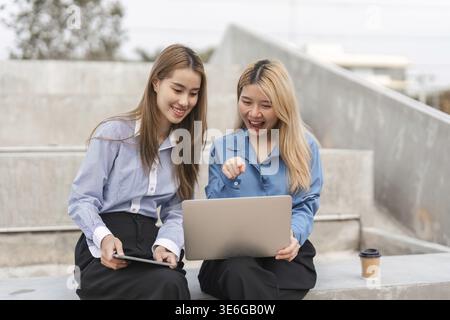 Two diverse women excitedly collaborating on a laptop and tablet, sharing ideas and insights in an outdoor setting Stock Photo