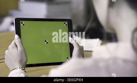 Rear view of a brunette woman using tablet with green screen, modern technologies concept. Female holding ipad with chromakey and reading something Stock Photo