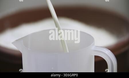 Pouring white liquid to a measuring cup with a container of flour on the background, cooking concept. Milk being poured in plastic measuring cup Stock Photo