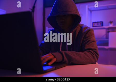 A mysterious hooded hacker working on a computer in a dark room. A concept for cybersecurity, hacking, cybercrime, and data privacy threats Stock Photo