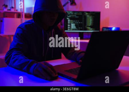 A mysterious hooded hacker working on a computer in a dark room. A concept for cybersecurity, hacking, cybercrime, and data privacy threats Stock Photo