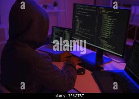 A mysterious hooded hacker working on a computer in a dark room. A concept for cybersecurity, hacking, cybercrime, and data privacy threats Stock Photo