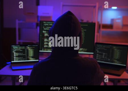 A mysterious hooded hacker working on a computer in a dark room. A concept for cybersecurity, hacking, cybercrime, and data privacy threats Stock Photo