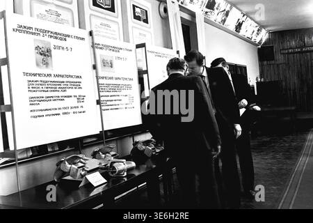 Sloviansk, 1983, AIZ plant exhibition room. Two male specialists in formal suits are closely examining massive metal armatures and supporting clamps presented on a table during an official product certification event. Large detailed technical posters detailing specifications and load capacities of the electrical hardware hang in the background. This authentic black and white archival photograph captures the rigorous engineering evaluation process of the early eighties and the everyday reality of a calm and peaceful Donbas. Stock Photo