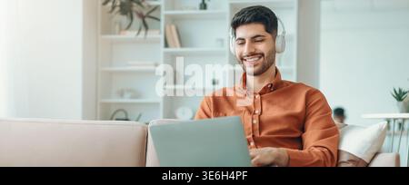 Young Man Works on Laptop in Cozy Living Room With Headphones on During the Daytime Stock Photo
