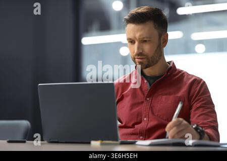 Man Working on Laptop and Taking Notes in Office Setting During Daytime Hours Stock Photo
