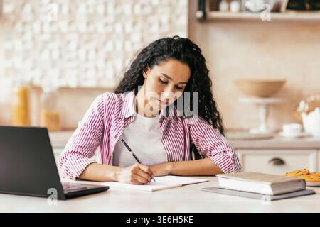 Woman Writes Notes at a Desk Near a Laptop While Sitting in a Kitchen Stock Photo