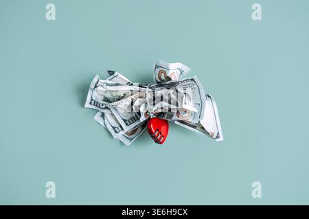 Crumpled hundred dollar bills are fastened with heart shaped combination lock on blue surface. Worry about money, financial stress concept, economic Stock Photo