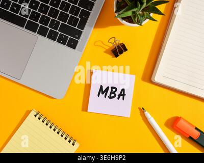 Office desktop with laptop, notebook, sticky note with MBA written on it. Master of business administration education concept. Stock Photo