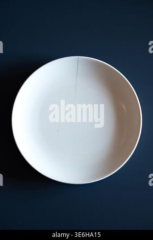 Minimalist top view of a white ceramic plate with a visible crack placed on a dark blue background Stock Photo