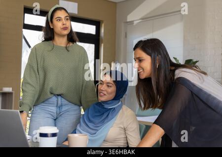 Diverse female coworkers collaborating around laptop in small office with coffee cups and hijab Stock Photo
