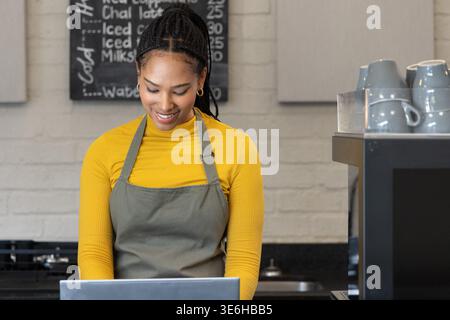 African American woman in gray apron smiling while typing on laptop at counter with coffee machine Stock Photo