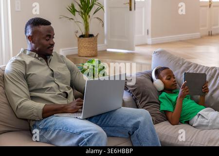African American father and son working on sofa, using silver laptop, tablet and white headphones Stock Photo