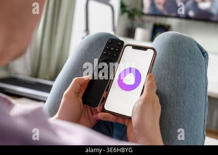 A man holds a smartphone with the Yandex Alice AI app and adjusts the TV remote. Saint Petersburg, Russia - 7 Apr 2026 Stock Photo