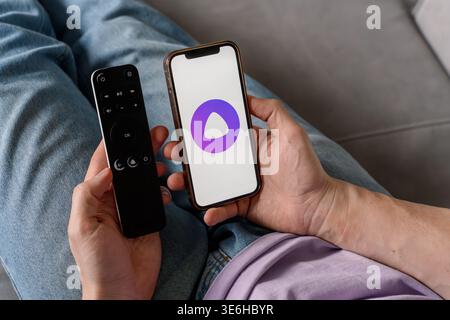A man holds a smartphone with the Yandex Alice AI app and adjusts the TV remote. Saint Petersburg, Russia - 7 Apr 2026 Stock Photo