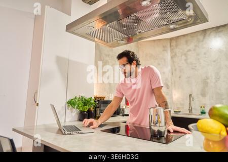 Man using laptop in modern kitchen, checking emails or working remotely while preparing coffee. Lifestyle of a young adult, freelancer Stock Photo