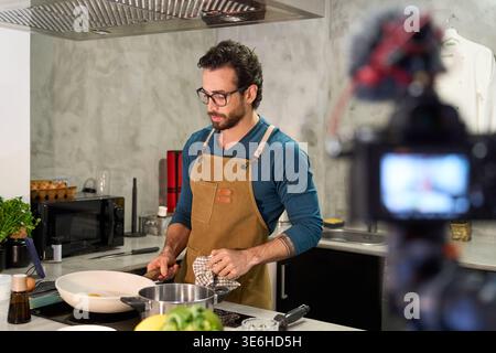 Man wearing an apron preparing food in a kitchen while recording a cooking tutorial for his online audience Stock Photo
