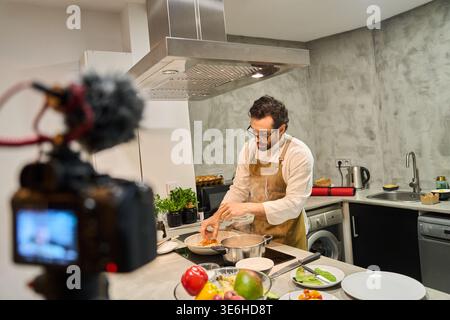 Chef recording a cooking tutorial in a modern home kitchen, filming a recipe video for social media with camera and microphone Stock Photo