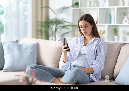 Disappointed woman in blue checking phone sitting on a couch at home Stock Photo