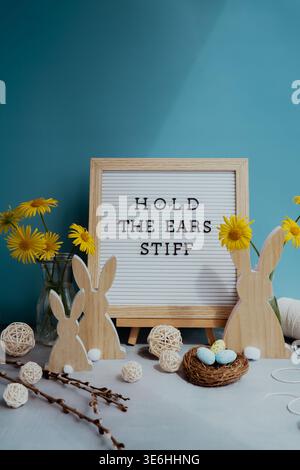 Phrase HOLD THE EARS STIFF traditional greeting to christian holiday. Festive mood composition with bunnies and yellow flowers on pastel blue background. Sustainable natural materials decorations Stock Photo