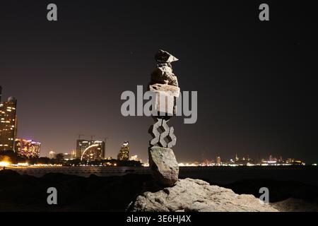 Zen tower made of stones, wooden pieces and paving blocks balanced on rock against blurred city lights and sea at night Stock Photo