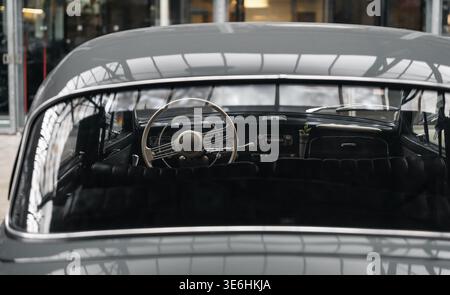 Interior view of a classic car through rear window. Old car interior with a thin ivory steering wheel, retro dashboard, and black leather seats. Stock Photo