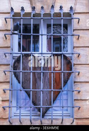 Decorative wooden heart placed behind window with metal bars on textured wall. Concept of love, protection and emotional distance. Soft natural light. Stock Photo