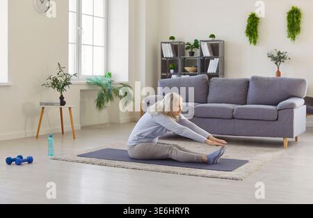 Senior elderly woman doing stretching exercises for health sitting on yoga mat at home Stock Photo