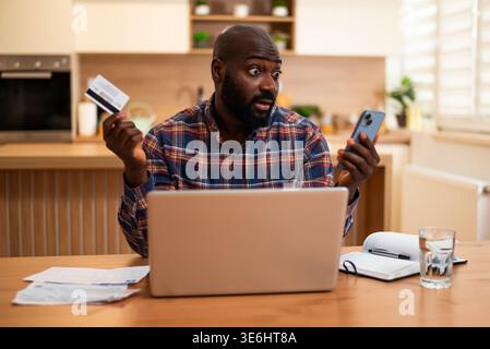 Surprised black man sits at home, holding credit card and smartphone, reviewing financial documents and calculating his expenses. Stock Photo