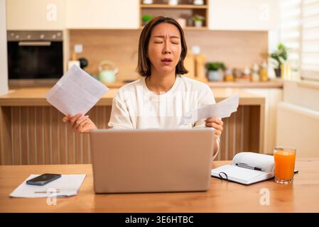 A worried woman sits at a table reviewing bills and documents while looking at her laptop, appearing stressed about her finances. Stock Photo