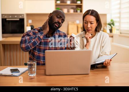 A couple sits at a table looking concerned while using a laptop, with the man holding his neck and the woman checking medication and taking notes. Stock Photo