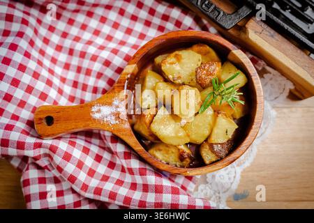 View of golden roasted potatoes nestled in a wooden bowl, sprinkled with salt and a sprig of rosemary, against a red and white checkered cloth, Pula, Stock Photo