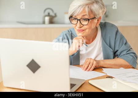 Online education courses webinar concept. Middle aged senior woman using laptop computer writing notes. Focused mature old woman enjoying studying online from home written records doing online work Stock Photo